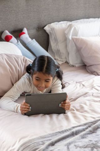 Asian girl lying on bed using tablet for learning and play, cozy pastel bedroom, home technology