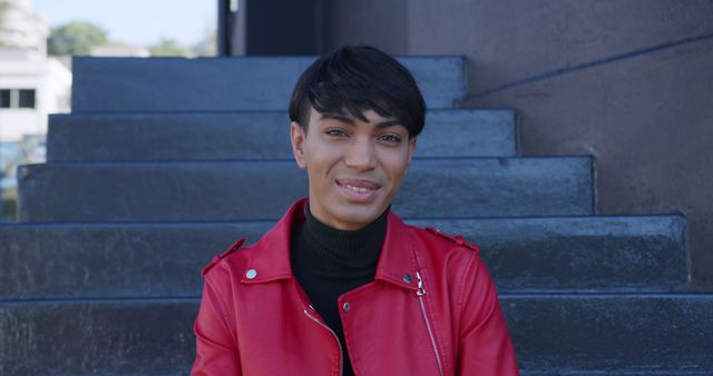 This image captures a young person smiling and sitting on outdoor steps, exuding confidence and style in a bright red jacket. Perfect for use in campaigns promoting youth lifestyle, fashion, urban settings, and streetwear brands. Great for editorial content, social media posts, and advertisements targeting a younger demographic. This image embodies a relaxed, trendy, and confident vibe.