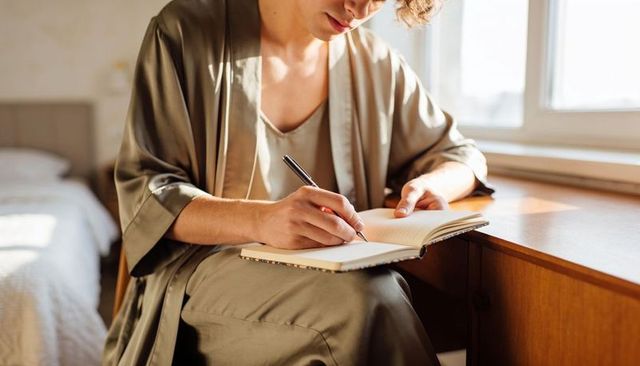Calm morning journaling woman wearing olive robe writing in notebook at sunlit desk