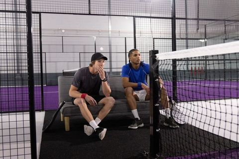 Diverse male athletes watching match sitting on bench at indoor padel court