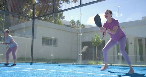 Mature women playing padel on sunlit blue home court with glass enclosure, doubles action