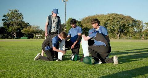 Youth cricket team preparing for practice with coach guiding players fastening pads