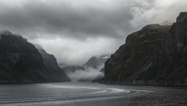 Dramatic misty fjord cutting between steep rocky cliffs with rippled wake and moody clouds