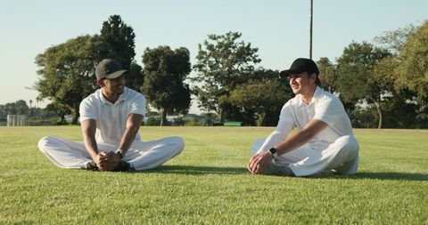 Diverse male teammates sitting and stretching on sunny sports field wearing white polos and caps