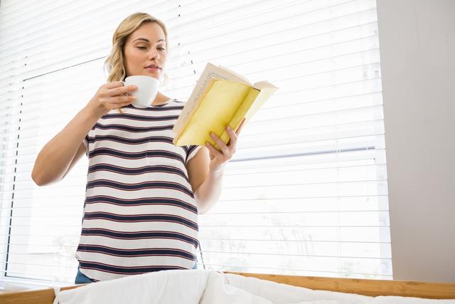 Woman standing by window, holding coffee cup and reading book. Ideal for lifestyle blogs, relaxation concepts, home comfort themes, and morning routines.