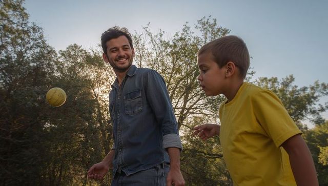 Smiling father playing catch with son in park at golden hour, tossing yellow ball