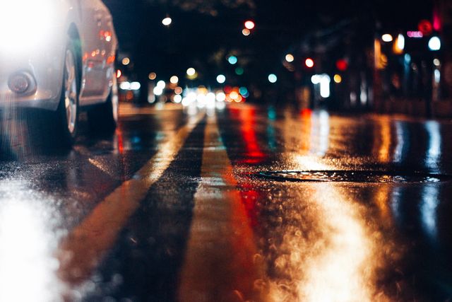 Low angle view capturing the vibrant reflections of city lights on a wet street at night. This image can be used for concepts like urban lifestyle, nighttime cityscape, rainy weather, and street photography.