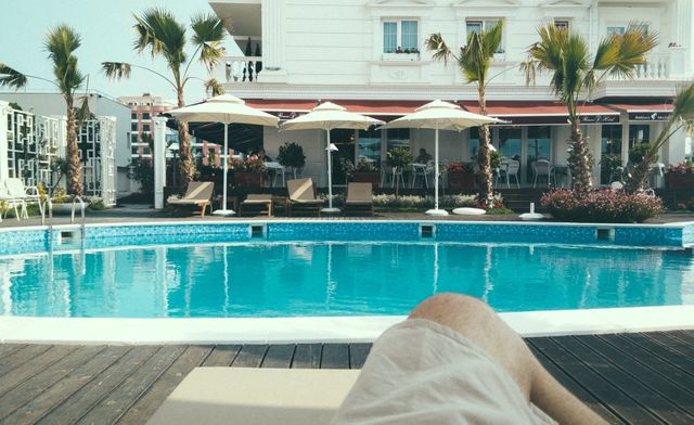 Person relaxing by the pool at a luxurious resort under sunny skies. Palm trees, comfortable loungers, and umbrellas provide shade, creating a perfect vacation atmosphere. Ideal for travel agencies, tourism promotions, resort advertisements, and summer vacation themes.