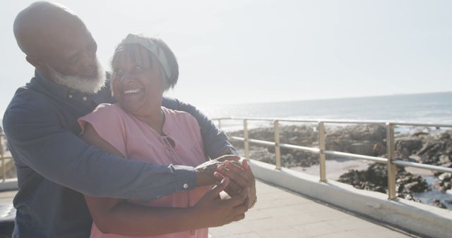Senior couple embracing on a sunny boardwalk by the ocean. Their joy and affection are evident as they smile at each other, wrapped in a loving hug. This can be used for themes related to relationships, retirement, vacation, coastal lifestyle, elderly well-being, and couples spending quality time together by the seaside.