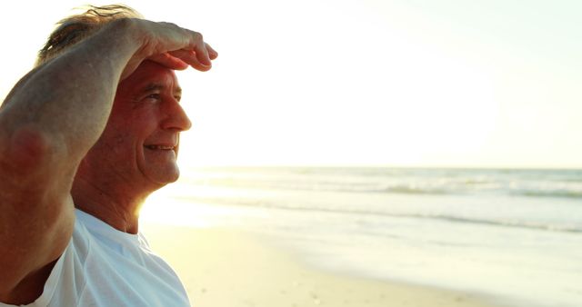 A middle-aged Caucasian man gazes into the distance on a sunny beach, shielding his eyes from the sun, with copy space. His expression suggests he is enjoying a moment of peace or searching for something in the horizon.