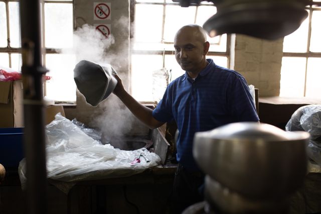 Front view of a bald biracial man holding fabric over a steam while creating a hat in the workshop at a hat factory.