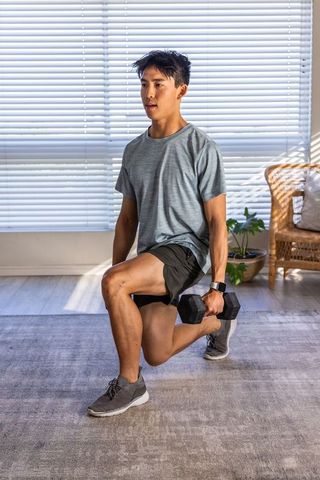 Asian man performing weighted lunge in sunlit living room wearing smartwatch