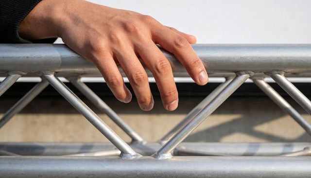 Right hand draping over aluminum truss railing close-up with diagonal shadows and texture