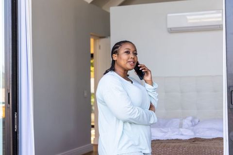 African american woman in blue pajamas talking on smartphone in minimalist bedroom