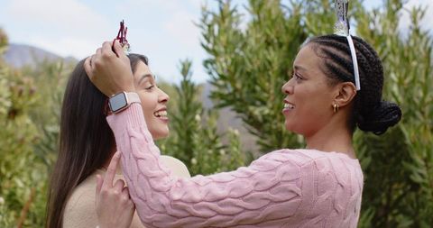 Multicultural friends placing tiara outdoors laughing and bonding in green garden wearing smartwatch