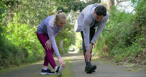 Diverse friends stretching hamstrings on leafy park trail wearing gear and wristwatch for run