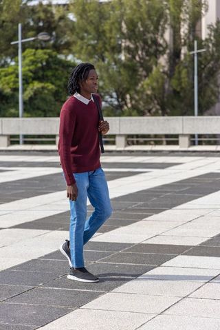 Young african american man walking across urban plaza wearing maroon sweater and carrying bag