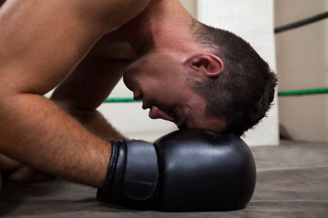 Young boxer resting with head on gloves in boxing ring. Ideal for use in sports, fitness, and motivational content, highlighting themes of perseverance, determination, and the physical demands of boxing.