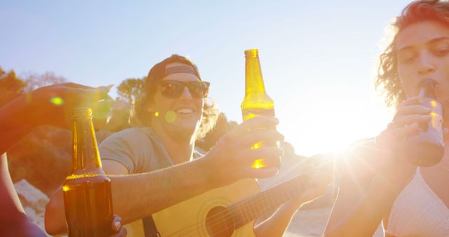 Group of friends enjoying bottled drinks and playing guitar at sunset on the beach. Perfect for depicting scenes of outdoor fun, socializing, summer leisure activities, and group enjoyment. Useful for marketing in travel, lifestyle, and alcohol beverage industries.