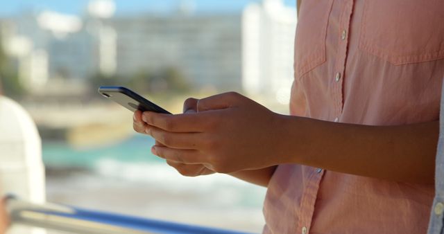 Woman texting on smartphone outdoors with blurred background of buildings and seafront water. Useful for images depicting communication, technology use, outdoor activities, social media engagement, and staying connected while on the move.