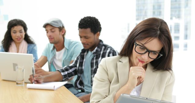 This stock photo depicts a diverse group of young professionals collaborating in a modern office environment. They are engaged with digital devices such as laptops and tablets, indicating a focus on technology and innovation. This image is ideal for use in articles or marketing materials related to teamwork, coworking spaces, remote work, digital transformation, and modern business environments.