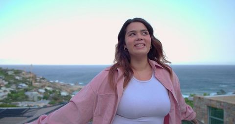 Young woman enjoying seaside view on sunlit balcony wearing pink shirt and white tank