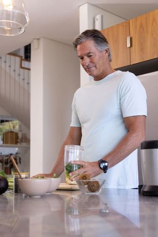 Mature man making green smoothie at modern kitchen island, chopping produce for blender