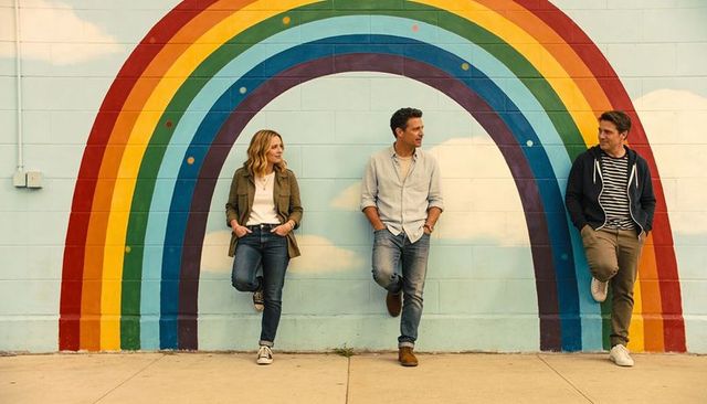 Three adults leaning on rainbow mural urban sidewalk chatting and laughing together
