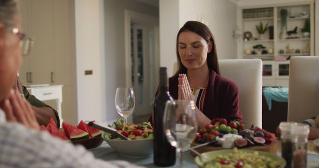 A woman with hands together praying while sitting at a dining table with family. Surrounded by a variety of dishes including fruits, vegetables, and wine, portraying a moment of gratitude and togetherness. Ideal for use in contexts related to family celebrations, traditional meals, thanksgiving, and expressions of faith.