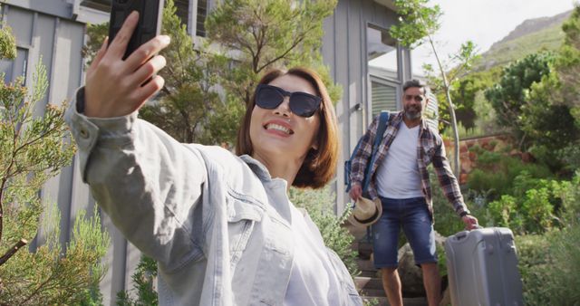 Couple arriving at vacation home, smiling and taking a selfie together. Man in background carrying luggage. Perfect for travel blogs, vacation rental promotions, holiday advertisements, and lifestyle content.