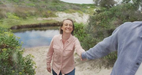 Smiling mature woman leading partner along coastal dune trail holding hand, candid outdoor
