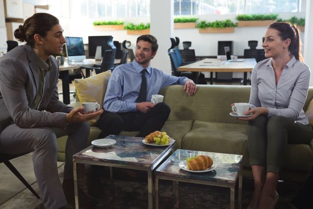 Executives are sitting on a couch in a modern office, engaging in a casual discussion while enjoying coffee and snacks. Ideal for use in articles about corporate culture, teamwork, business meetings, and professional environments.