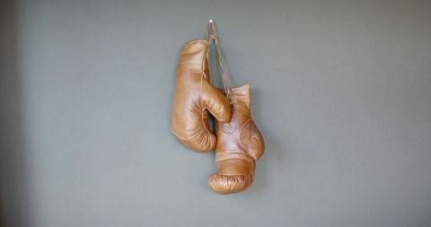 Vintage tan leather boxing gloves hanging by laces on gray wall with worn patina
