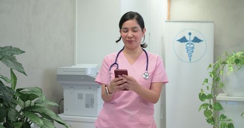 Nurse in pink scrubs standing and texting on smartphone with stethoscope in clinic