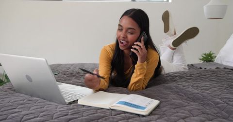 Young woman working from bed, talking on phone and pointing pen at laptop
