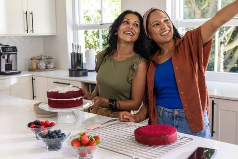Friends smiling and taking selfie while decorating red velvet cake on sunlit kitchen