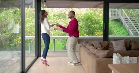 Young couple dancing in sunlit living room by glass doors with green backyard view
