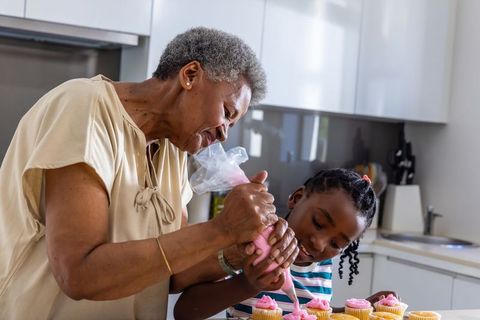 Senior african woman and girl decorating pink frosted cupcakes together in bright kitchen