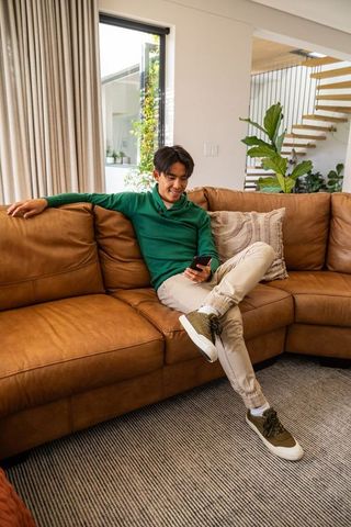 Asian man relaxing on brown leather sectional sofa in modern living room using smartphone