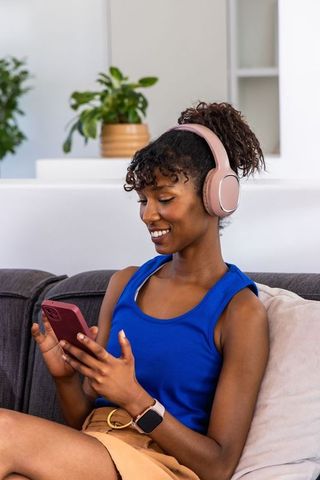 African american woman wearing pink headphones while scrolling smartphone and relaxing on couch