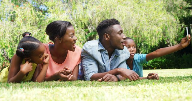 African American family laying on grass and taking a selfie. Ideal for use in advertisements promoting family time, outdoor activities, or technology. Useful for brochures, social media, and lifestyle blogs focused on togetherness, happiness, and family bonding.