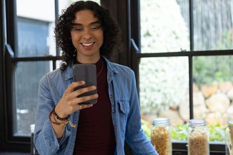 Non-binary person smiling at smartphone while standing at sunlit kitchen counter with pasta jars