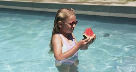 Young girl enjoying watermelon while wading in backyard pool wearing heart sunglasses