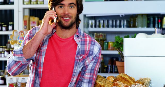 A cheerful man in a flannel shirt is talking on a mobile phone while standing in a bakery. He is surrounded by various types of bread and pastries. Shelves in the background are stocked with jars, bottles, and grocery items. This image is ideal for use in projects related to customer service, small businesses, bakeries, lifestyle blogs, and communication.