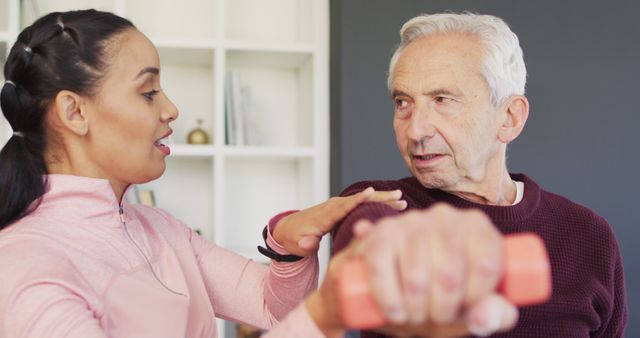 Senior man is receiving exercise guidance from a personal trainer holding a dumbbell at home. Useful for promoting physical therapy, elderly fitness programs, at-home rehabilitation services, and active lifestyles for seniors.