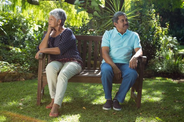 Senior couple sitting on a wooden bench in a lush green park, facing away from each other, indicating a disagreement or argument. The image can be used to depict relationship issues, communication problems, or emotional tension among elderly couples. Suitable for articles, blogs, or advertisements related to senior relationships, counseling, or mental health.