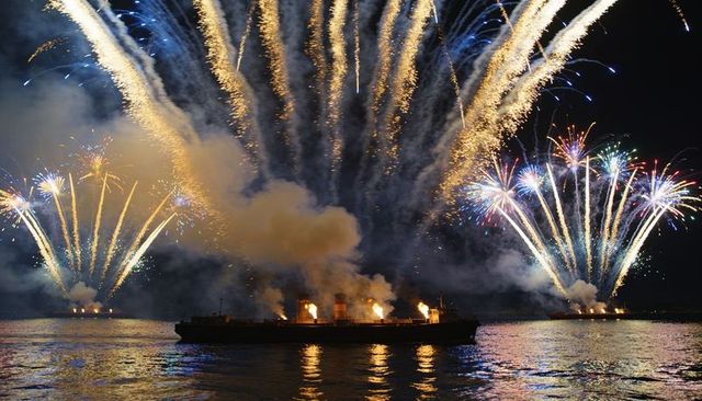 Silhouetted barge amid spectacular harbor fireworks, deck flames and reflective water