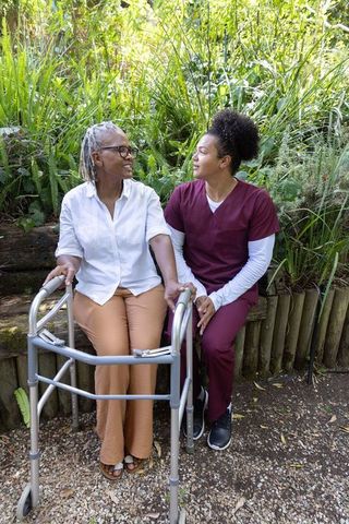 African-american senior woman holding hands with caregiver in sunny garden sitting with walker