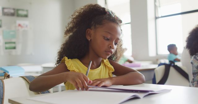 This image of a young girl concentrating while writing in a classroom can be used for educational resources, school promotions, and children's learning materials. Perfect for illustrating concepts of focus, learning, and childhood education.