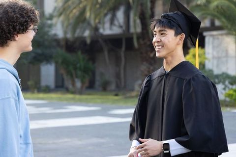 Smiling graduate in cap and gown celebrating with friend on university campus walkway
